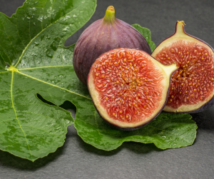 "Close-up of two whole figs and one halved fig with vibrant red interiors and purplish skins, resting on large green fig leaves with water droplets, set against a dark textured background."